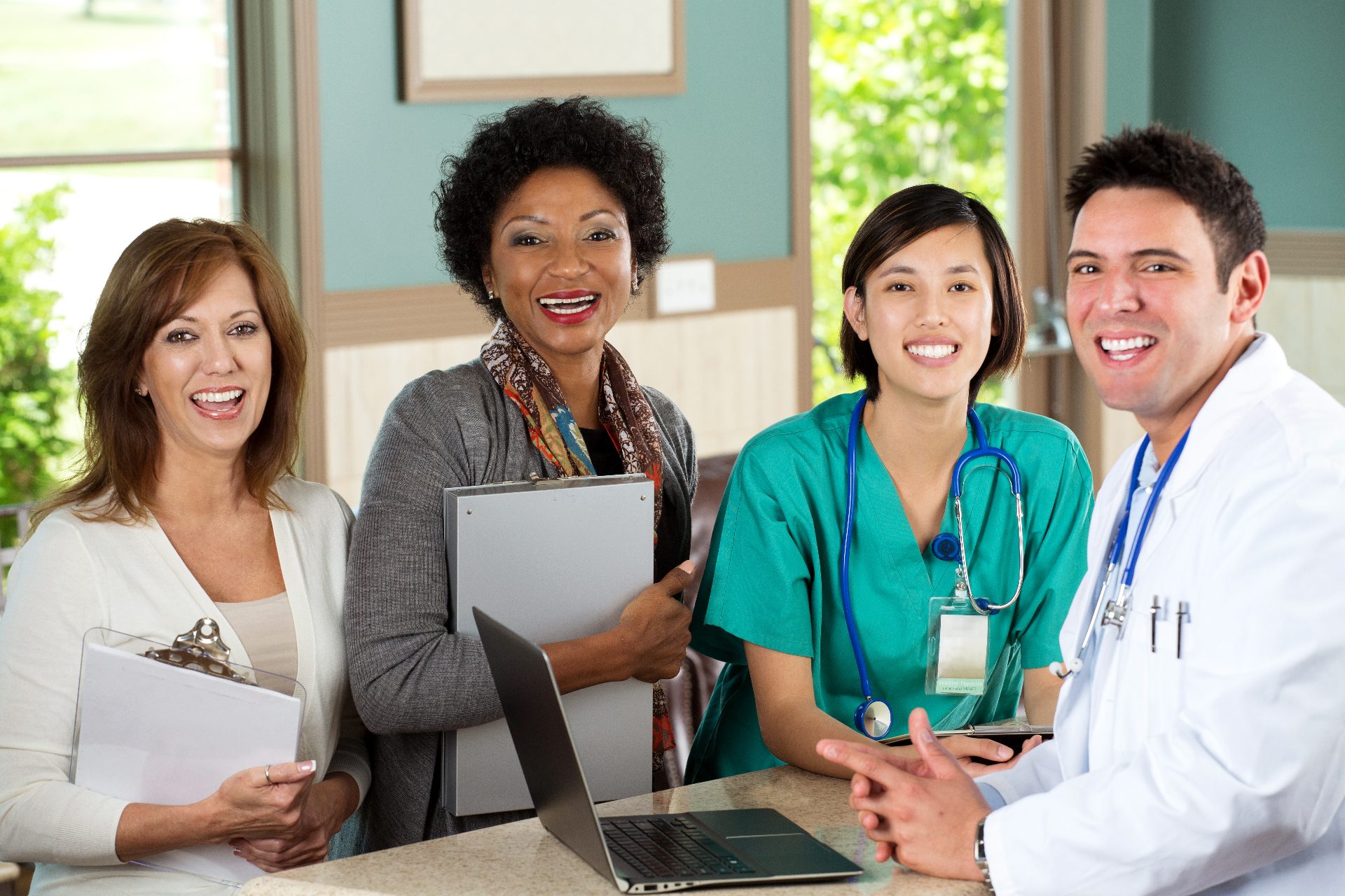 Four smiling medical office staff at a front counter with clipboards, a laptop and a stethoscope
