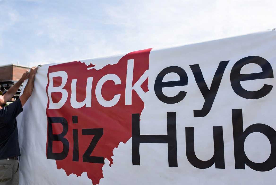 Person hanging a large white vinyl banner with the Buckeye Biz Hub logo outdoors