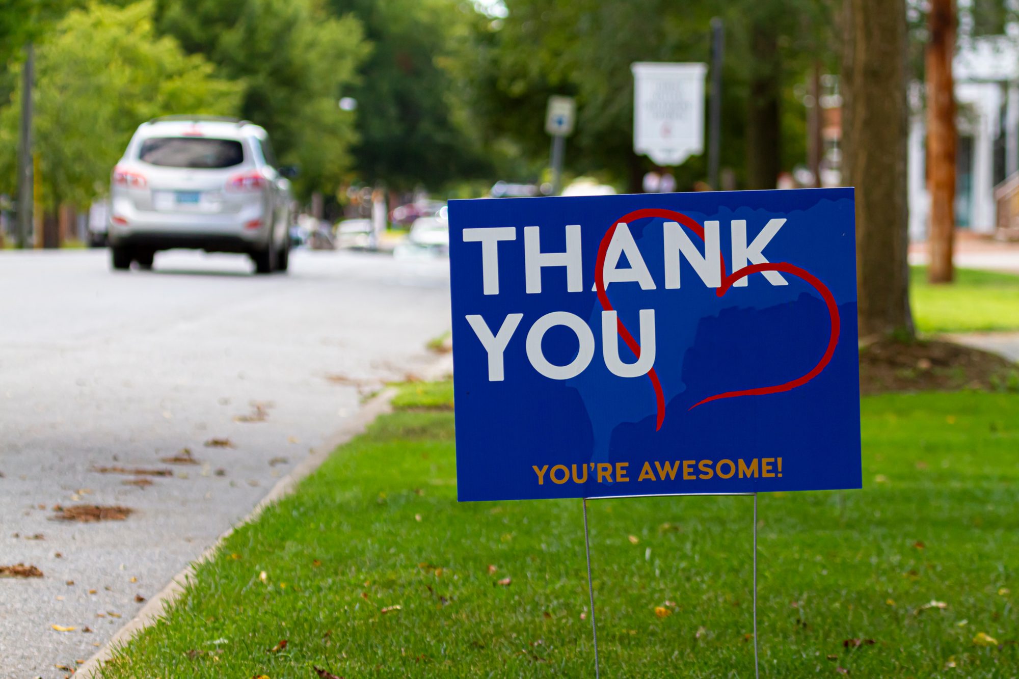 Blue thank-you yard sign on wire stakes in the grass beside a tree-lined street